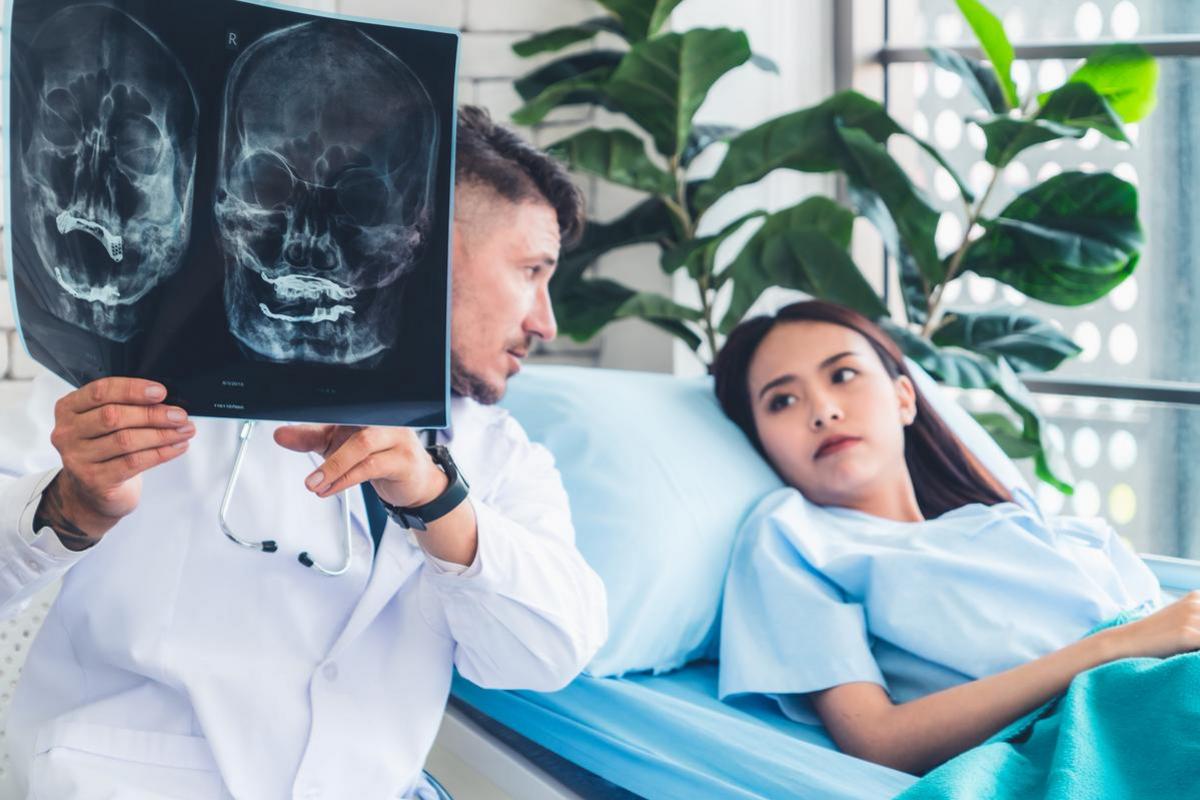A clinician holds skull x-rays beside a young woman resting on a hospital bed.