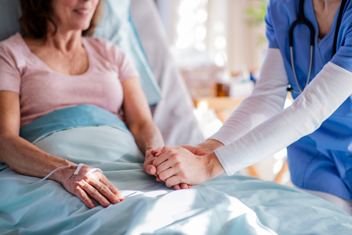A clinician in blue scrubs holding the hand of a smiling patient resting in a hospital bed